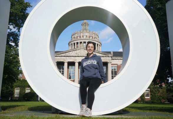 Sarah Woodams sitting in the O of the Meliora sign with Rush Rhees in the background