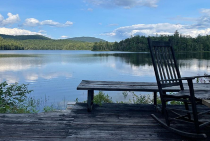 photo of a lake under a blue sky with mountains in the background and a small dock and rocking chair in the foreground.