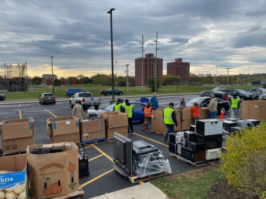 boxes of electronics, volunteers helping people empty cards. Campus and sky in the background.